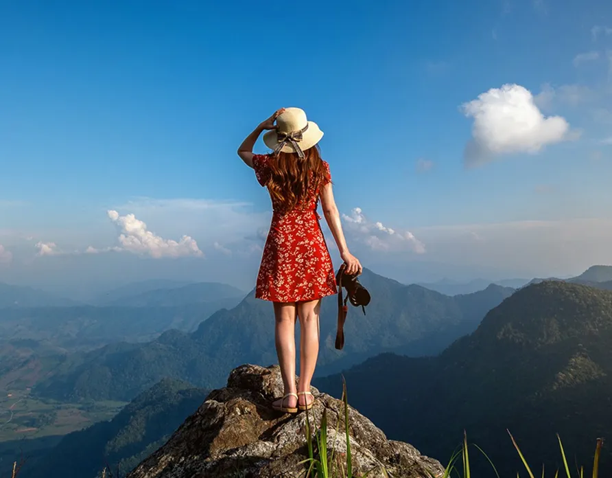 Woman in a red dress and sunhat standing on a rocky peak overlooking mountains