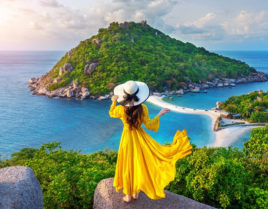 Woman in a flowing yellow dress and large hat on a rock overlooking a green island and blue sea