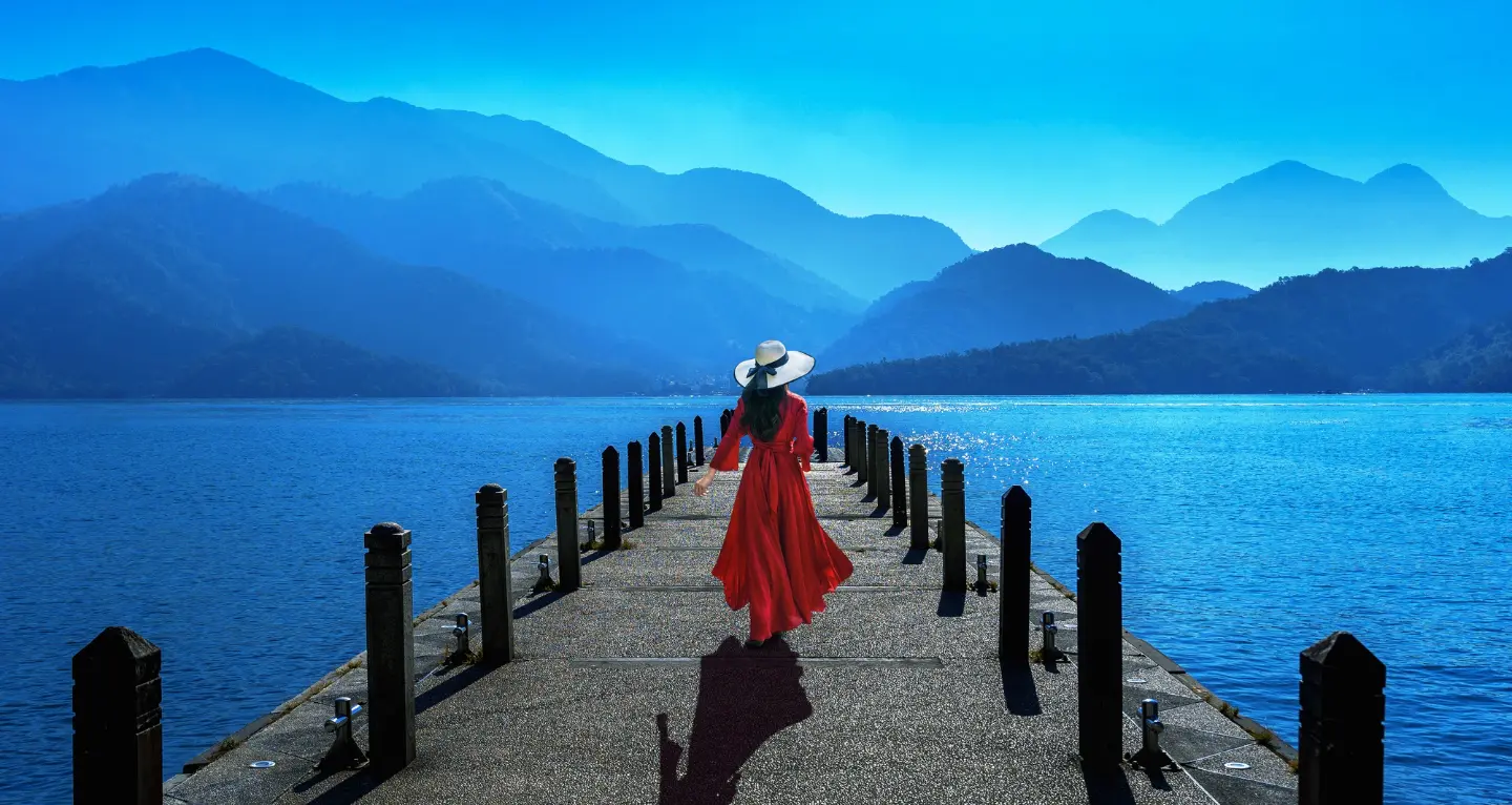 Woman in a red dress and white hat walking on a pier toward a blue lake